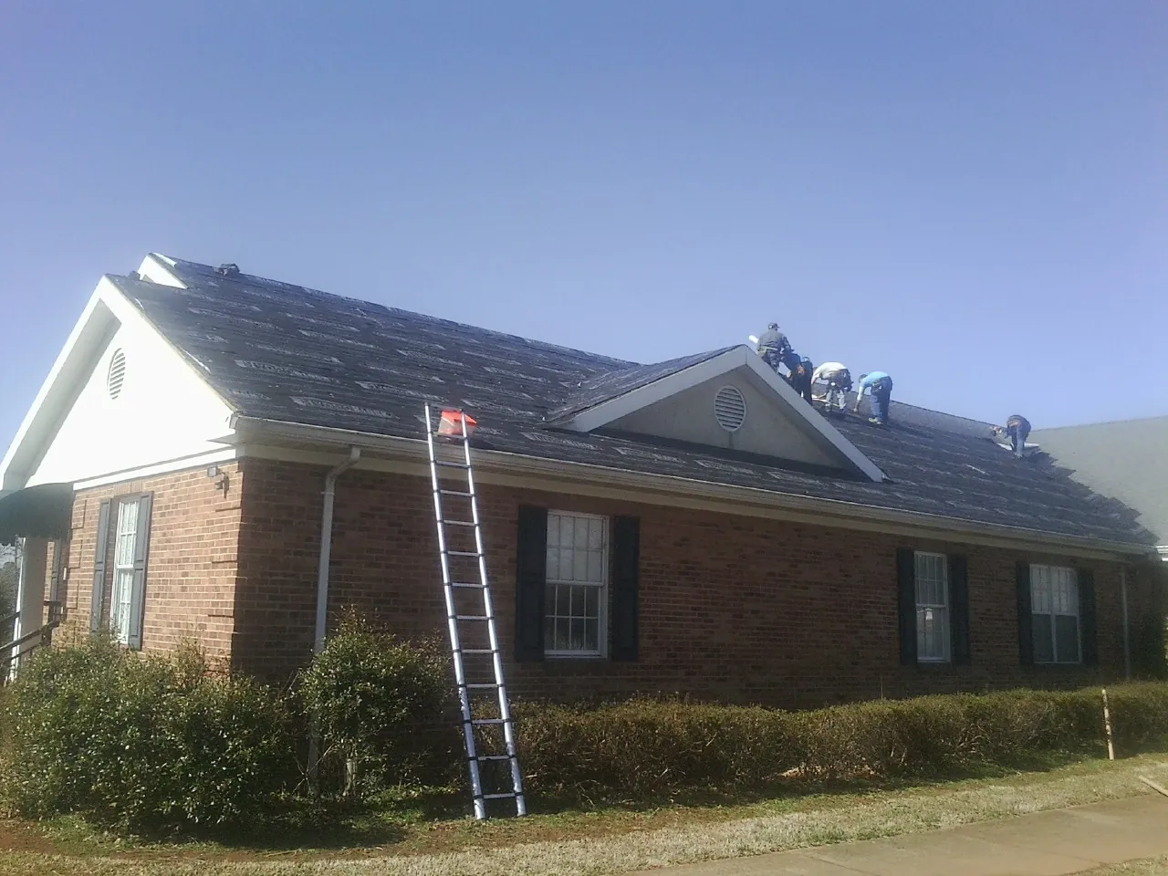 Crew tearing shingles off a brick ranch roof with a ladder set — Emergency Roof Repair work by our crew in Ludowici