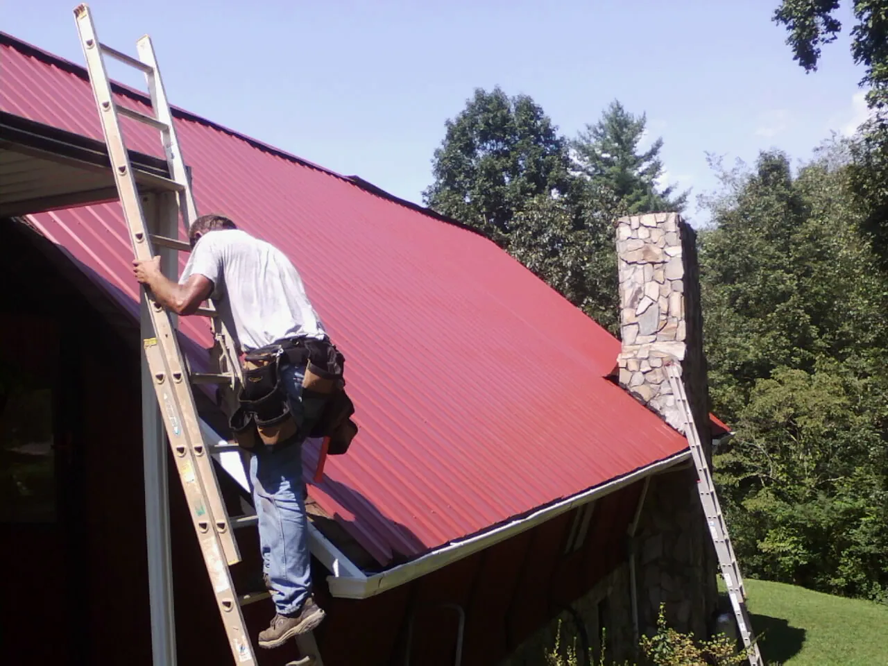 Roofer on a ladder beside a red metal roof and stone chimney — Soffit & Fascia Repair by Roofing Brothers in Ludowici